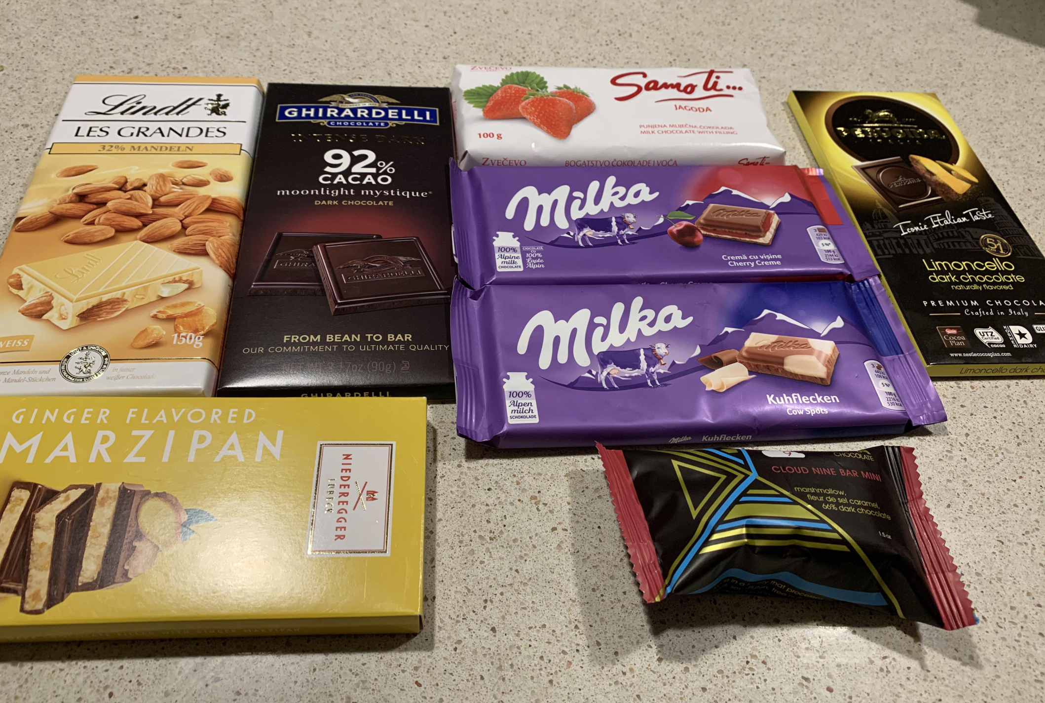 An assortment of chocolate bars and sweets, including Lindt, Ghirardelli, Sang Lin, Ferrero, Marzipan, Milka, and Urocello, arranged on a light-colored countertop.
