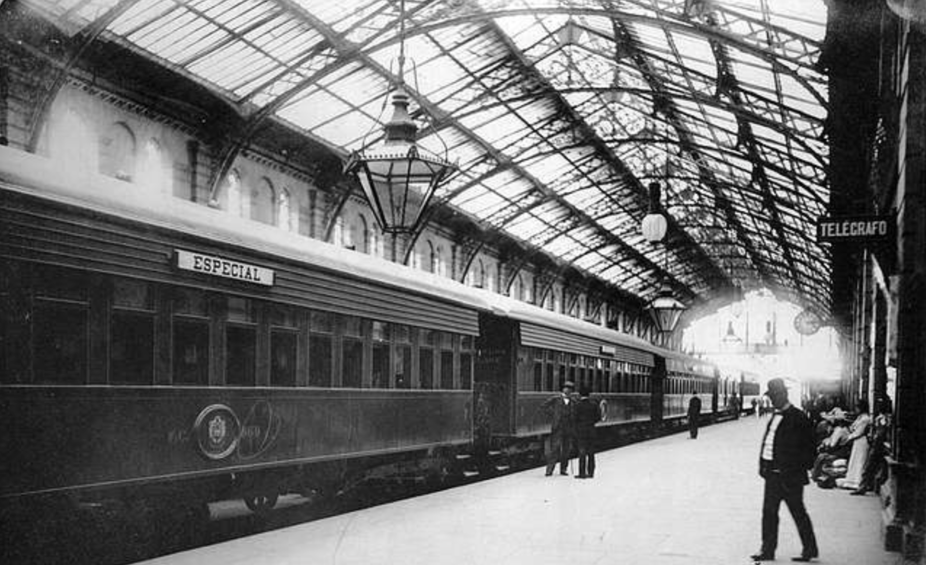 Black and white photo of a vintage train labeled "ESPECIAL" at a covered train station platform. People stand and sit along the platform beneath a glass and metal roof. A sign reads "TELEGRÁFO.