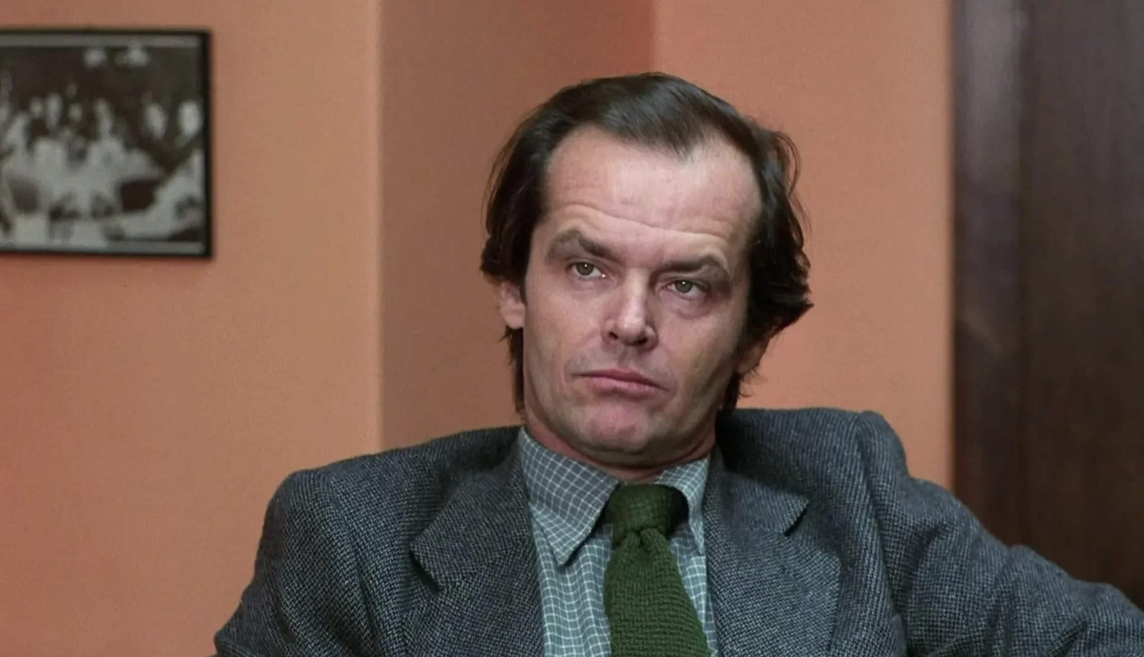 A man in a gray suit, checkered shirt, and green tie sits indoors against peach-colored walls, looking serious with a neutral expression. A black-and-white photo is visible on the wall behind him.