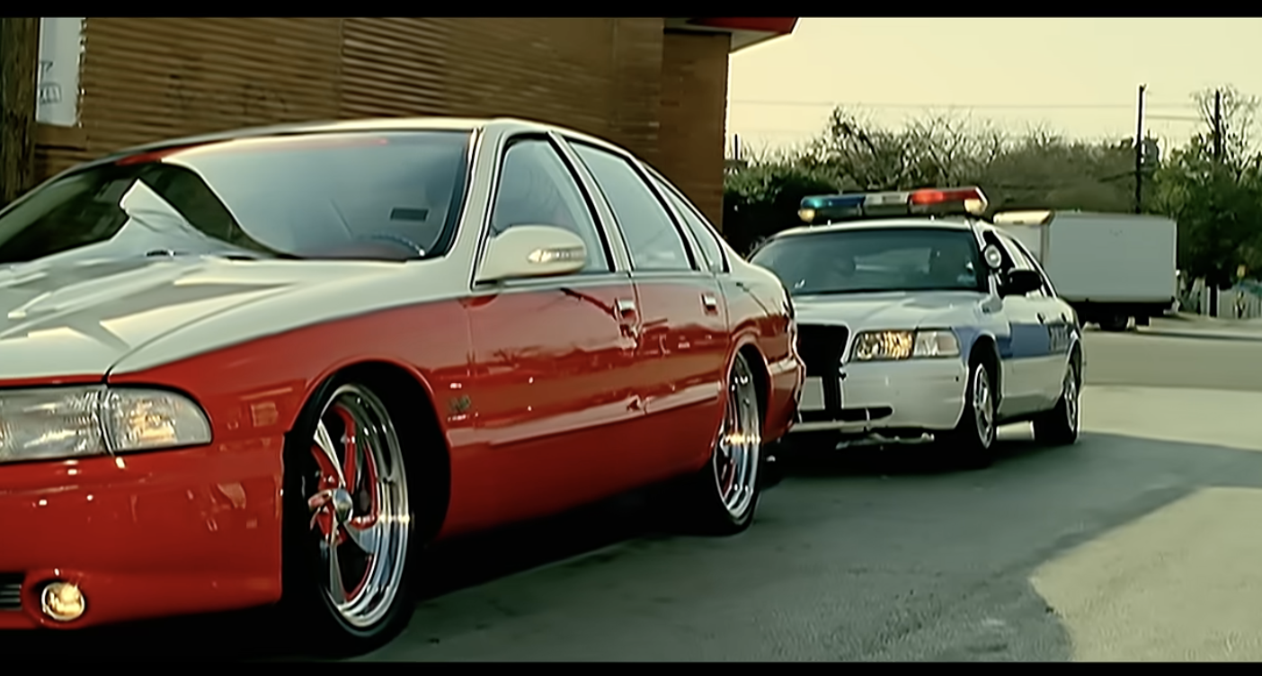 A red and white car with custom wheels is pulled over at the side of the road by a police car with flashing lights, in front of a building and trees.