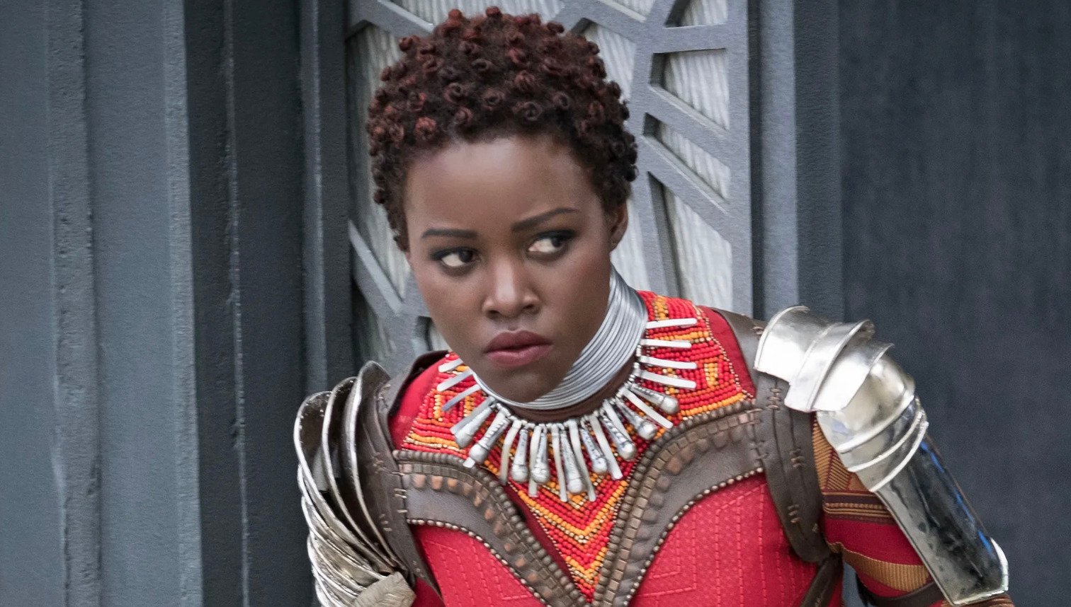 A woman in elaborate red and gold armor with a beaded necklace stands alert, looking to the side. She has short curly hair and is positioned in front of a patterned gray wall.