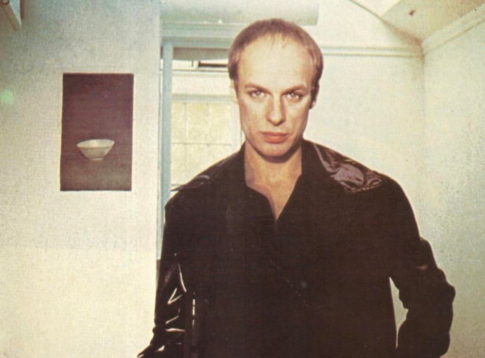 A person with short, light hair and a serious expression stands indoors, wearing a black leather jacket. There is a painting of a bowl on the wall behind them and soft natural light coming from a window.