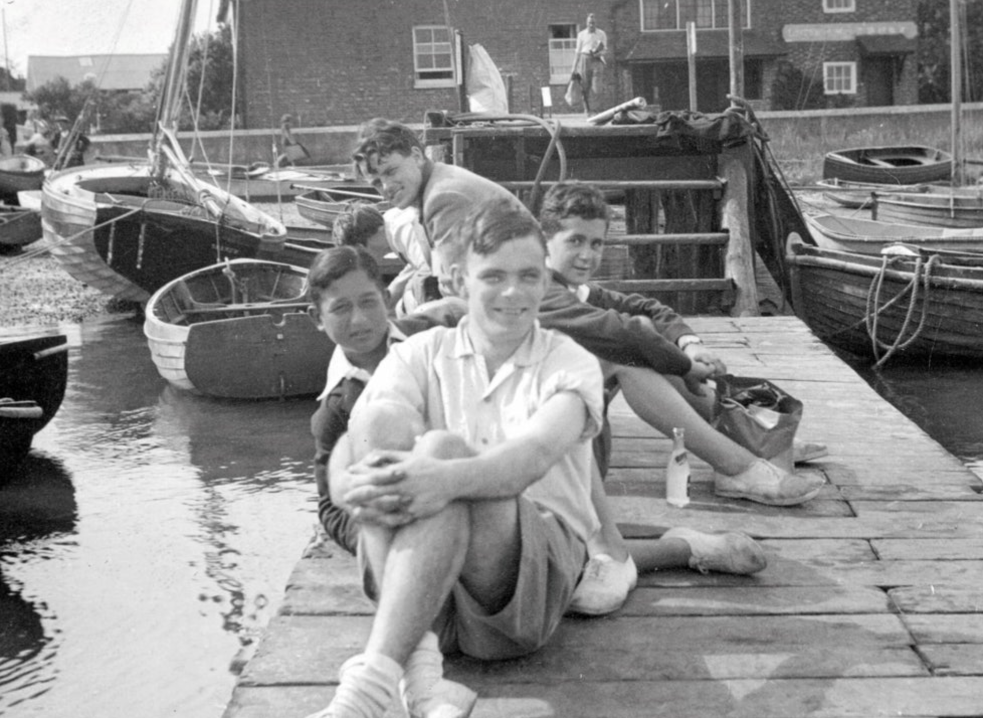 Four young people sit closely together on a wooden dock by the water, with boats moored beside them. They are smiling and casually dressed, appearing relaxed on a sunny day. Houses are visible in the background.