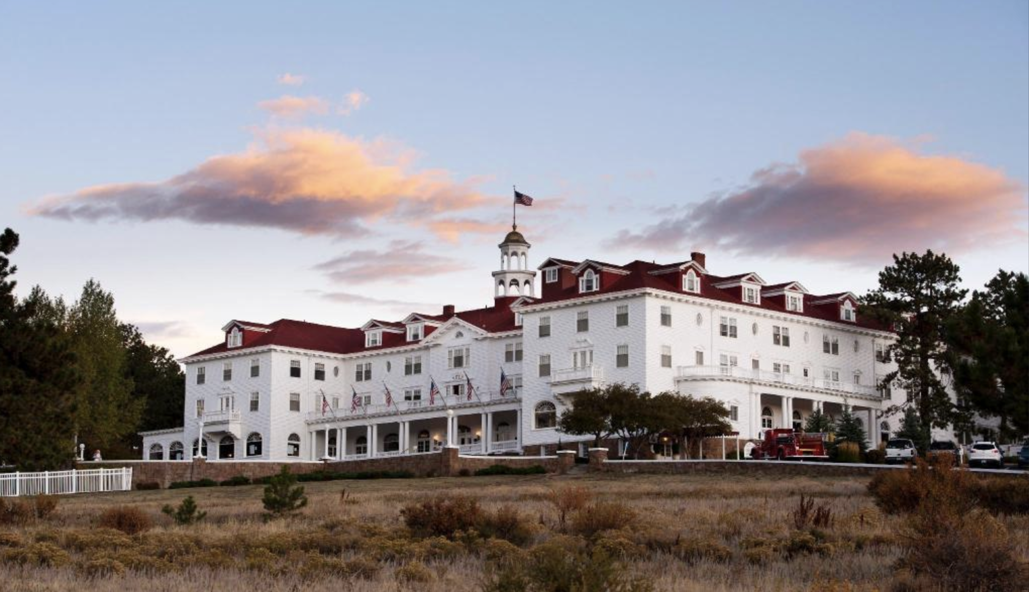 A large white historic hotel with a red roof and central cupola, surrounded by trees and grass under a sky with pink clouds at sunset. An American flag flies atop the building.