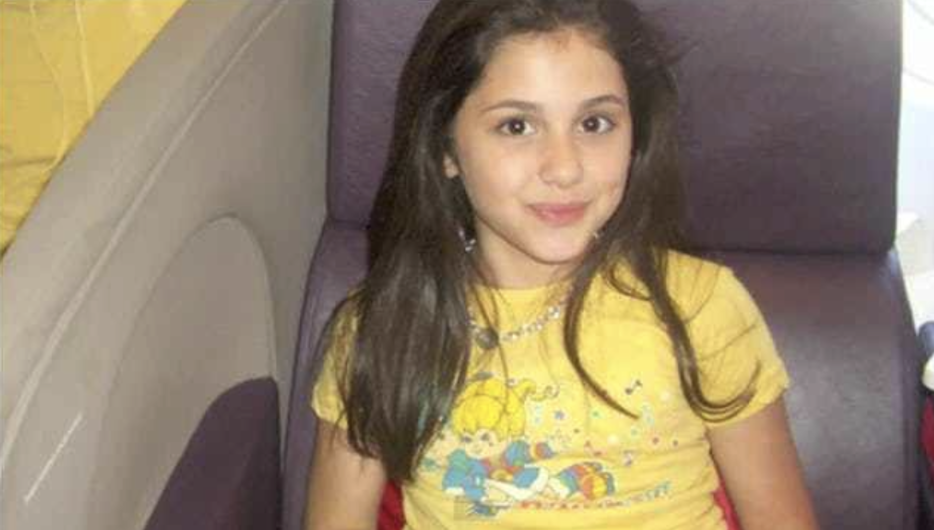 A young girl with long dark hair sits on a purple seat, smiling slightly. She is wearing a yellow T-shirt with a colorful cartoon print. The background includes part of a curved wall or seat.