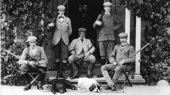 Five men in early 20th-century hunting attire pose with rifles on a house porch, two seated with three standing behind them. Two dogs lie in front of the group. The porch and building are covered with ivy.