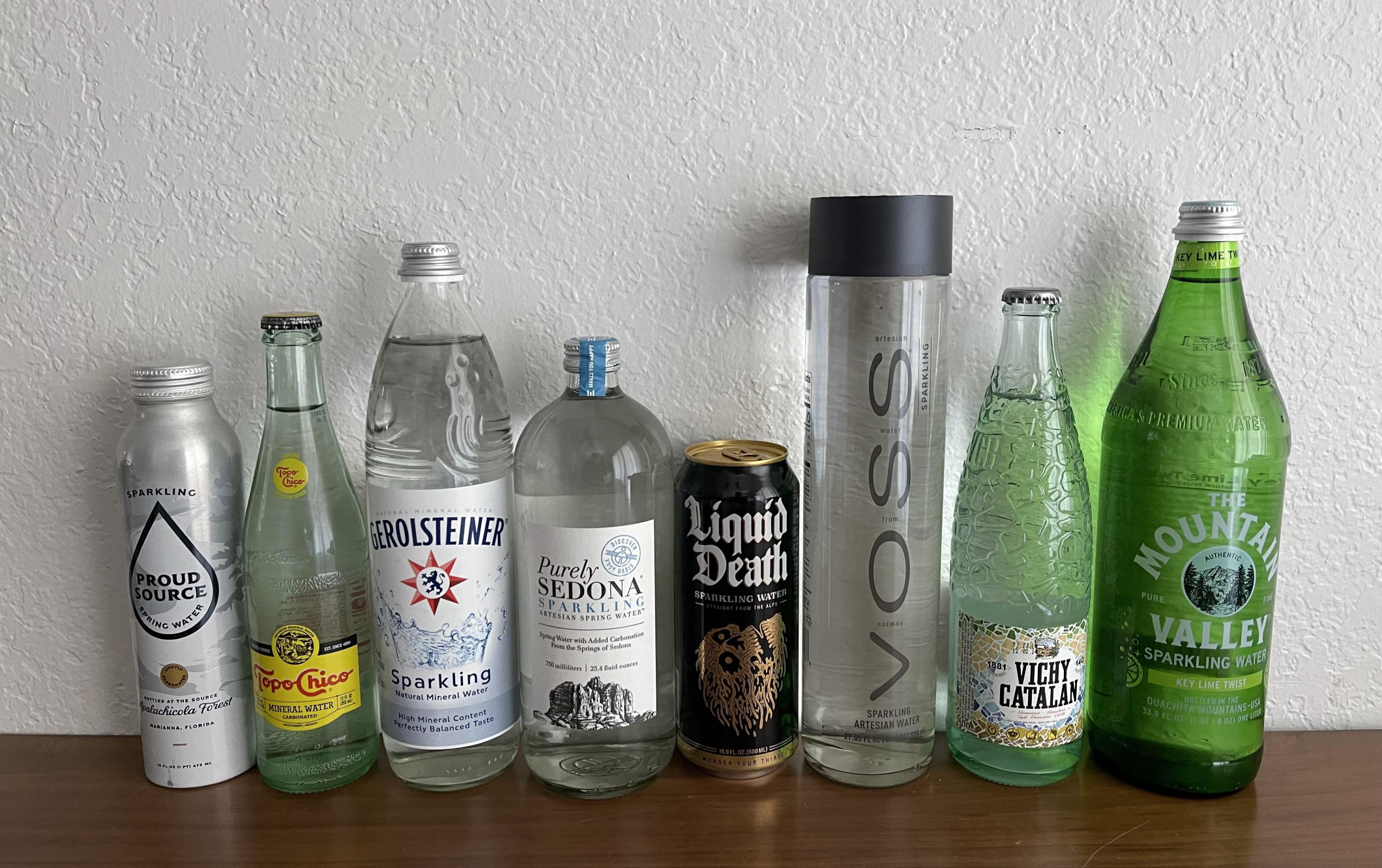 Eight bottles and cans of sparkling water from different brands, lined up on a wooden surface against a white wall. The containers are various shapes, sizes, and colors, some clear and others metallic or green.