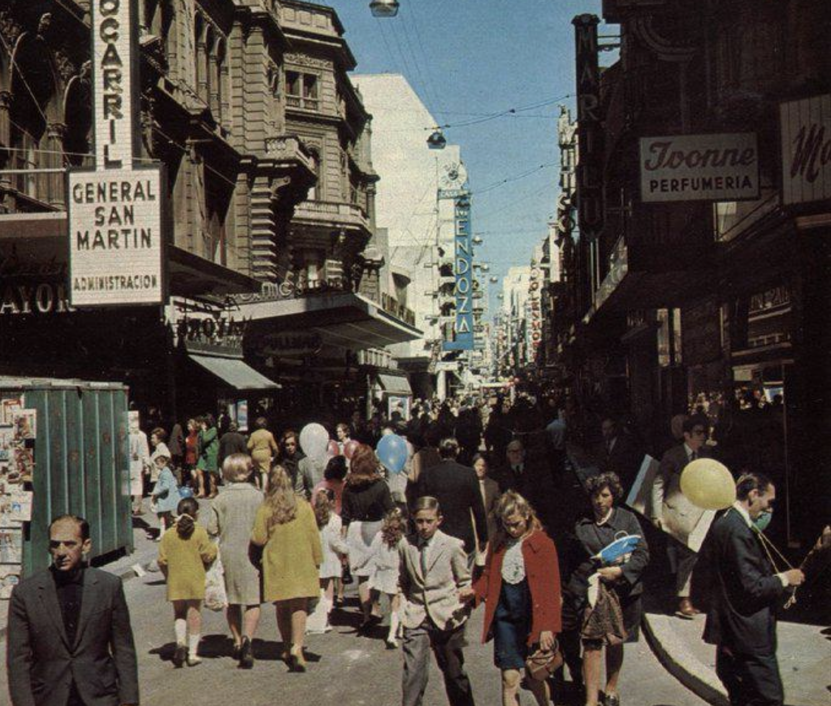 A busy city street scene from the past, with many people walking and shopping. Some carry balloons; storefront signs and classic architecture line both sides. The atmosphere is lively and bustling.