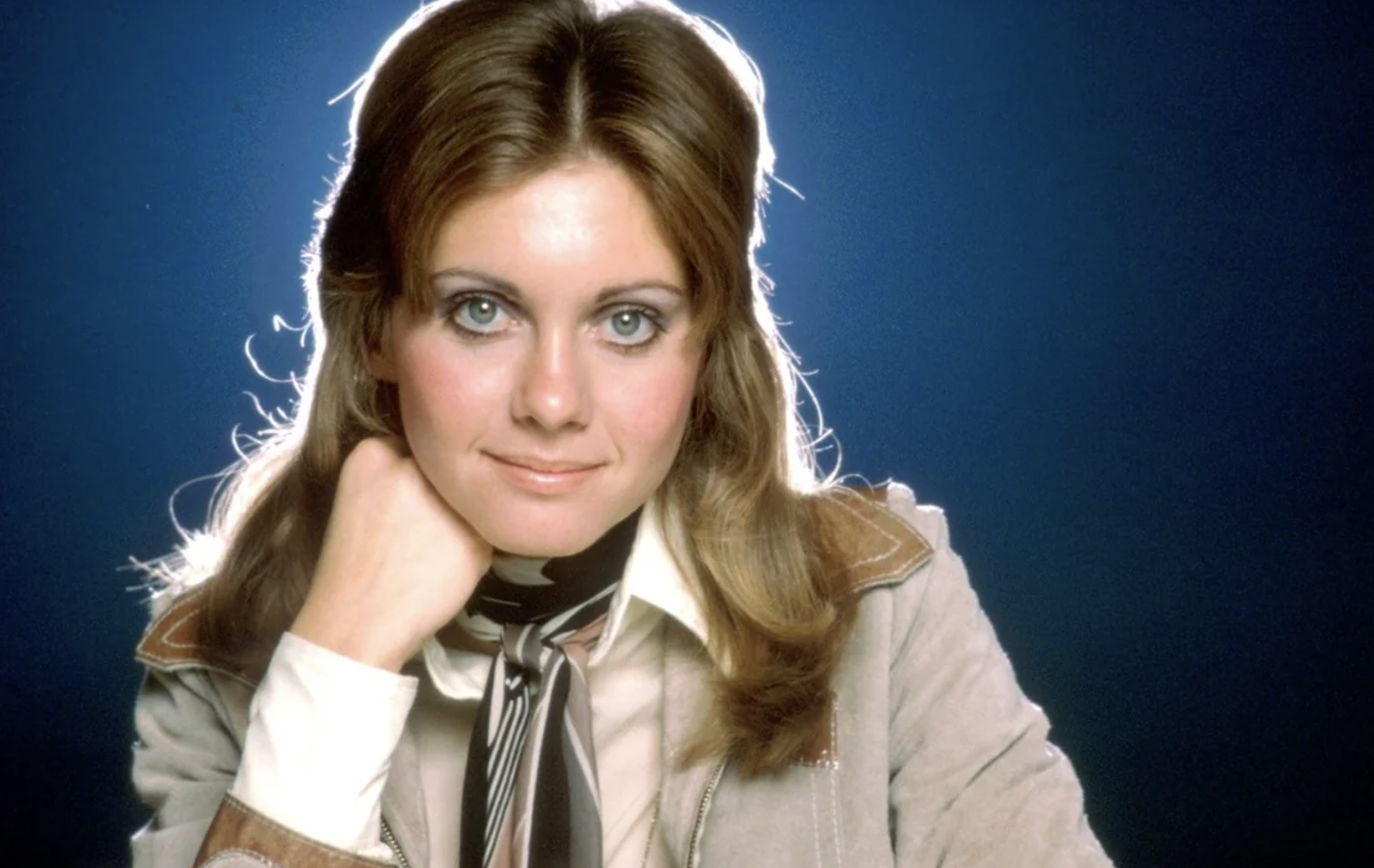 A woman with light brown hair, blue eyes, and a slight smile rests her chin on her hand. She wears a beige jacket, white shirt, and striped scarf, posing against a dark blue background with a glowing outline around her head.
