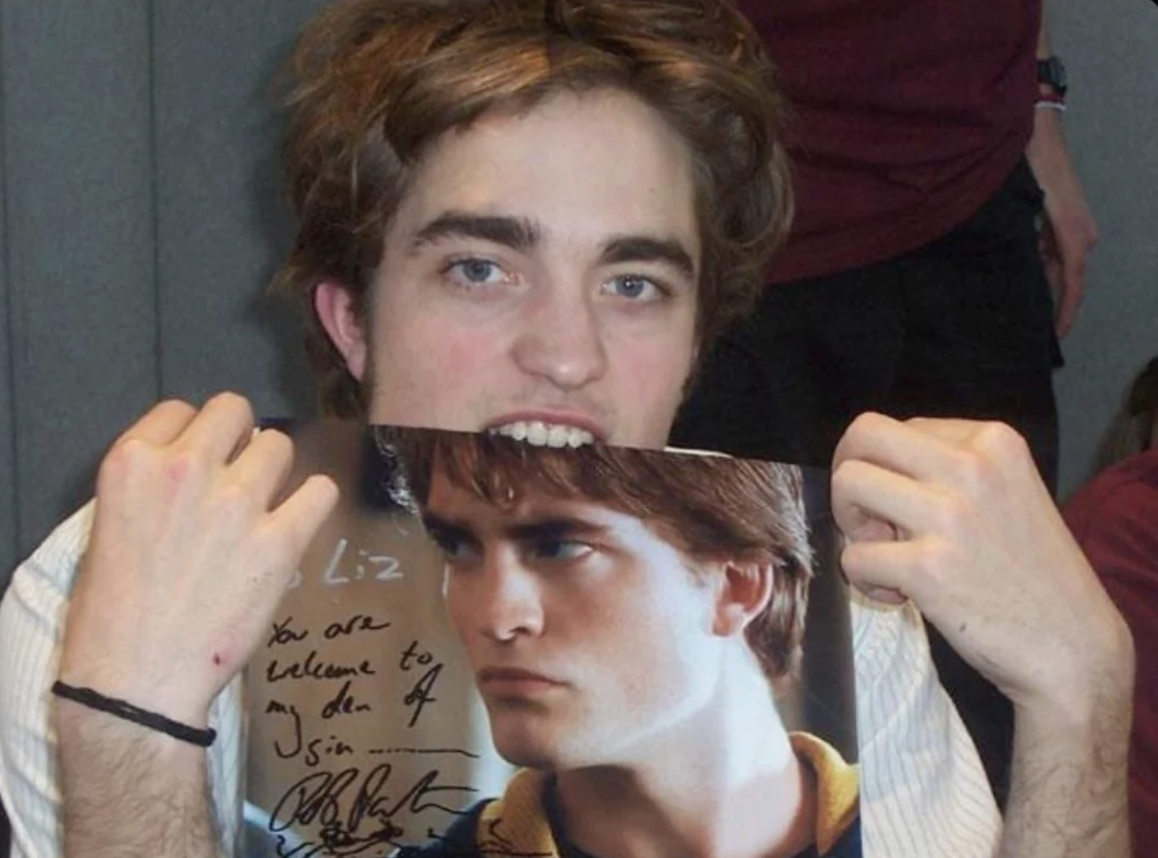 A young man playfully pretends to bite a signed photo of himself as a movie character, holding it up to his mouth with a grin, mimicking the vampire role he portrays in the image.