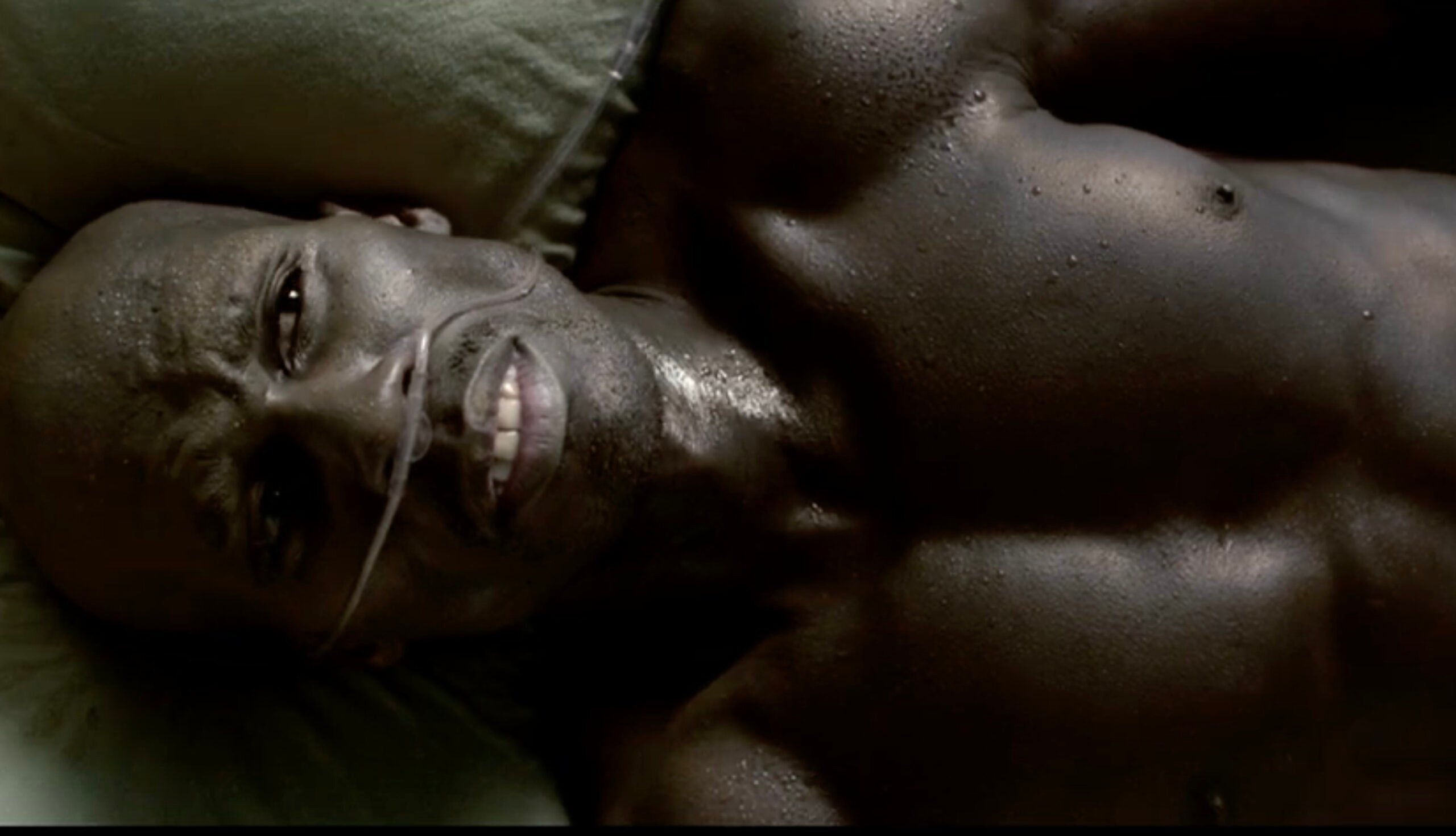 A shirtless man lies on his back, sweating and grimacing in pain, with an oxygen tube under his nose. The lighting emphasizes the tension in his expression and muscles.
