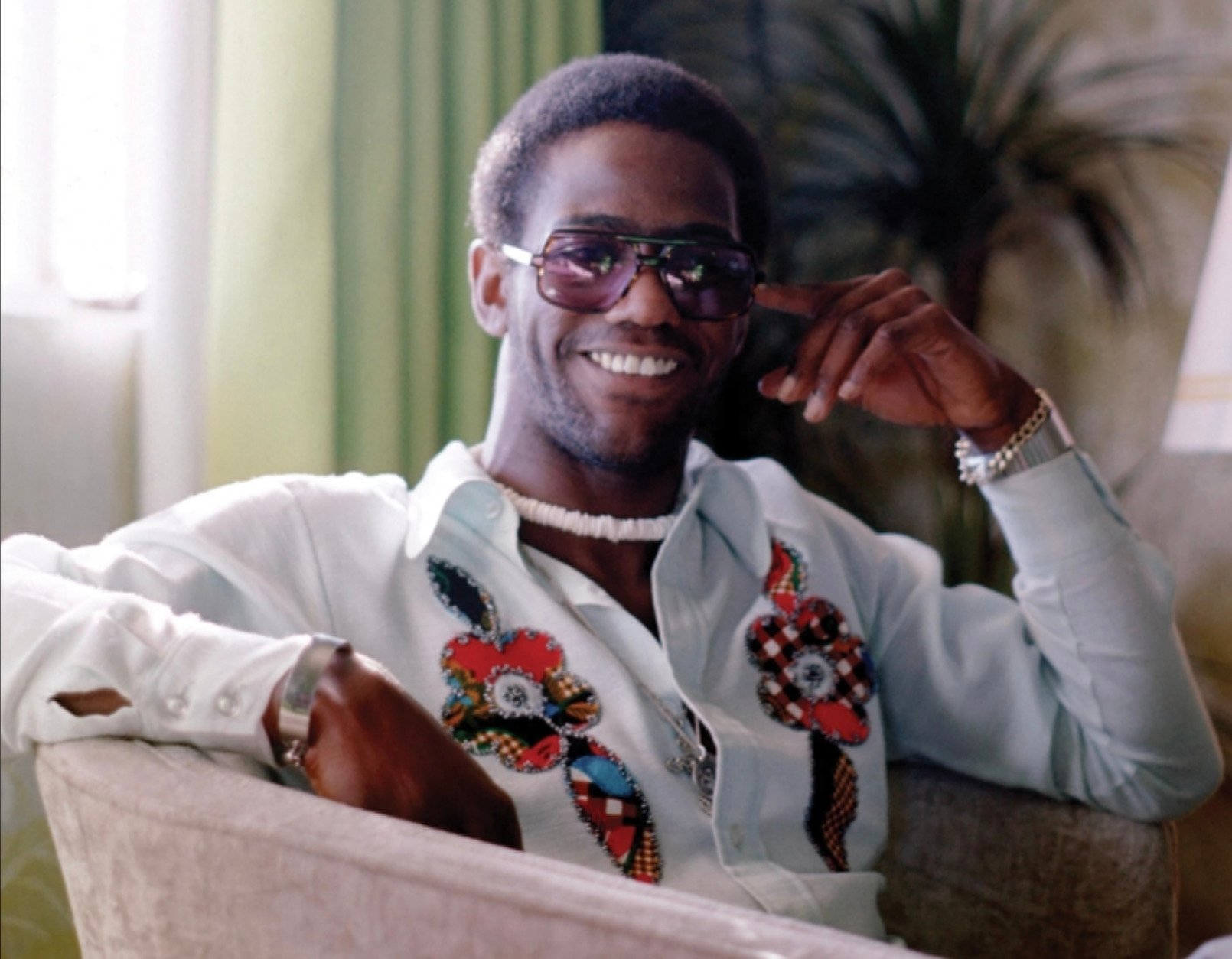A man wearing sunglasses, a beaded necklace, and a white shirt with colorful floral designs smiles while sitting on a light-colored couch. There is a plant and window with green curtains in the background.