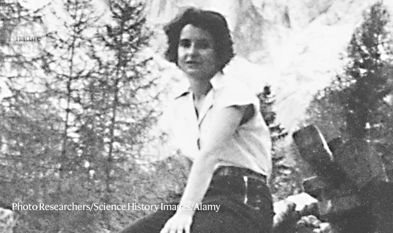 A black-and-white photo of a young woman with short hair, wearing a short-sleeve shirt and pants, sitting outdoors near trees and mountains. The image includes a watermark and a photo credit at the bottom.