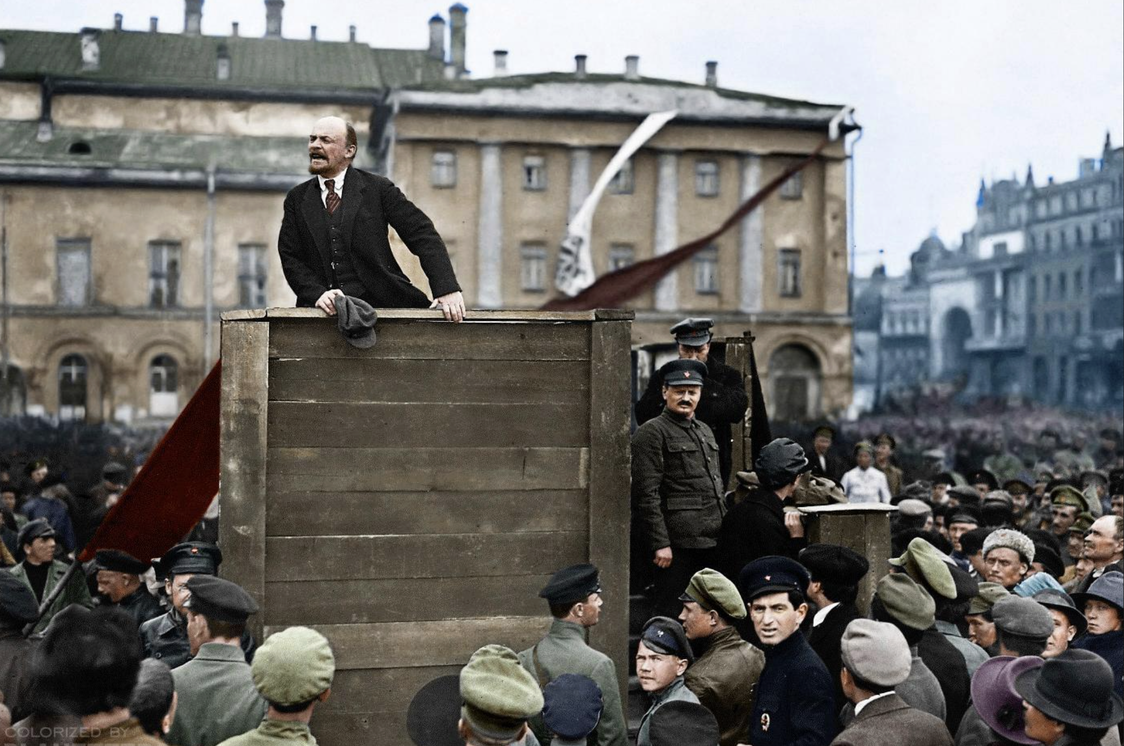 A man stands on a wooden platform speaking to a large crowd in a city square, surrounded by people in early 20th-century clothing, with historic buildings in the background.