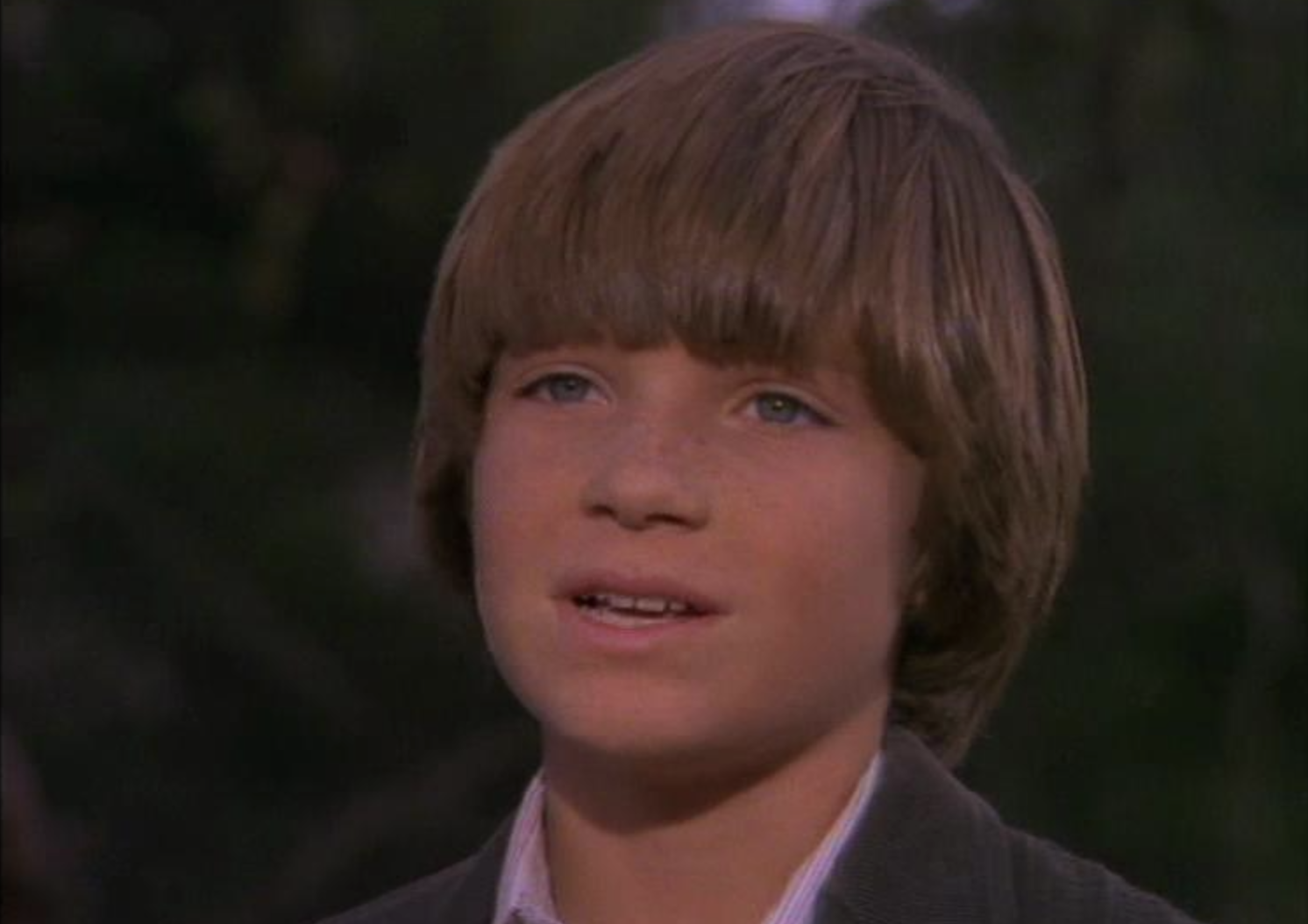 A young boy with light brown hair and blue eyes is outdoors, wearing a brown jacket and looking slightly to the side with a neutral expression. The background is blurred greenery.