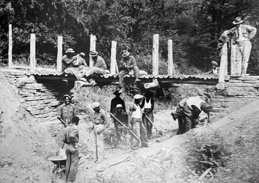 Black and white photo of men constructing a small log bridge; some stand or sit on the bridge, while others below dig and work with shovels and wheelbarrows on a sloped dirt path.