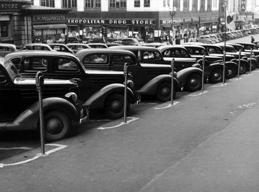 Black and white photo of several vintage cars parked in a row at parking meters on a city street, with storefronts including a drug store visible in the background.