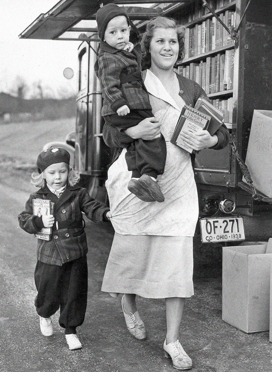 A smiling woman in an apron holds several books and carries a young child, while an older child walks beside her holding a book. They are in front of a bookmobile with shelves of books and an Ohio license plate dated 1938.