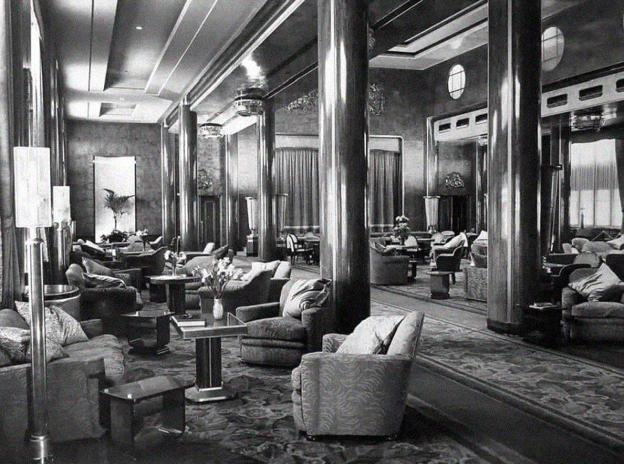 Elegant black-and-white photo of a spacious, luxurious lounge with plush armchairs, ornate carpets, tall, shiny columns, chandeliers, and large windows with curtains, evoking vintage 1930s or 1940s interior design.