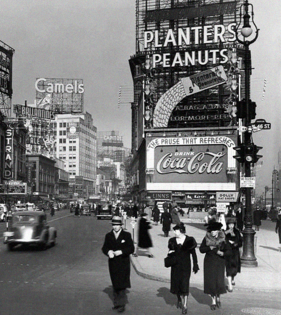 Black-and-white photo of a busy city street in the 1940s with vintage cars, pedestrians, and large advertising signs for brands like Planters Peanuts, Camels, and Coca-Cola.