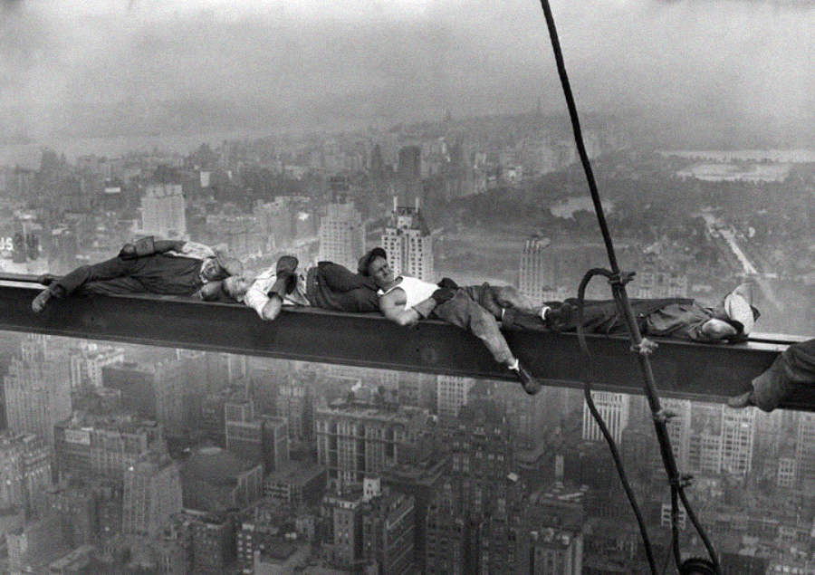 Four construction workers lie down and rest on a steel beam high above a city skyline, with buildings and trees visible below in the background. The scene appears foggy and the workers are relaxed despite the height.