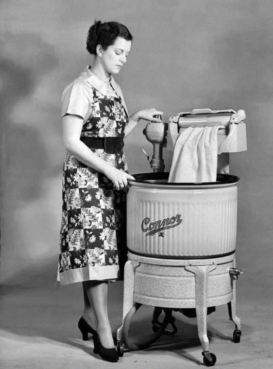 A woman wearing a floral dress and apron stands beside a vintage Conner washing machine, feeding laundry through a wringer. The image is in black and white, evoking a mid-20th-century setting.
