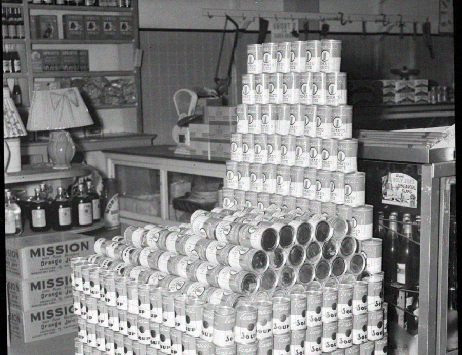 A black and white photo of a grocery store shows cans of Campbell's soup stacked to form a chair shape. Shelves with various products, a counter, and glass bottles are visible in the background.