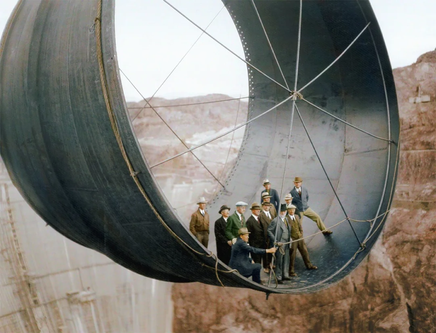 A group of workers in hard hats stand inside a large, suspended steel pipe high above the ground, with rocky cliffs and construction far below.