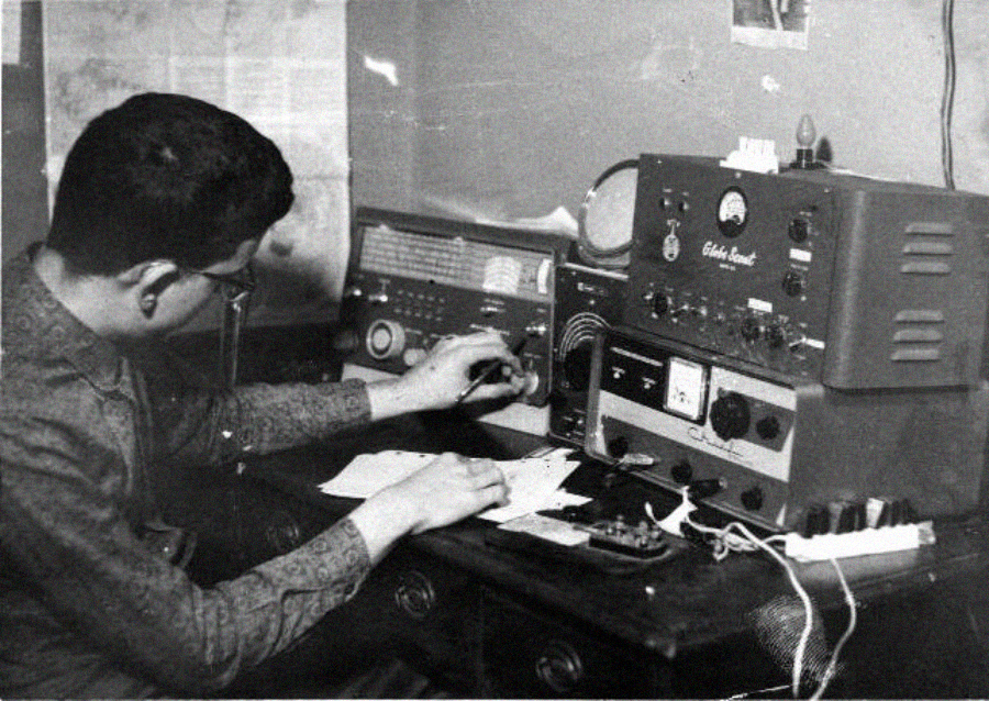 A person sits at a desk operating vintage radio equipment, adjusting dials and writing on paper, with several radios, meters, and cables visible on the desk in a black-and-white photo.