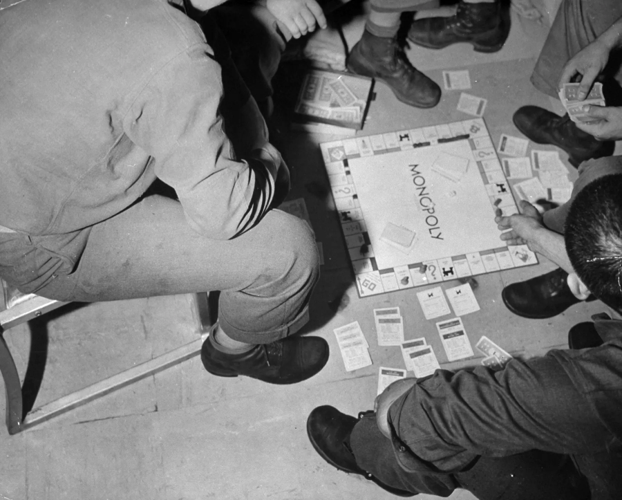 Four people sit on the floor, playing a game of Monopoly. The board and game cards are scattered around them, and only their lower bodies and hands are visible. The focus is on the game in progress.