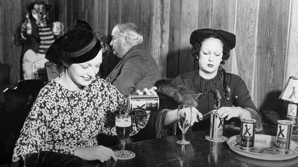 Two women in vintage clothing sit at a wooden table, pouring canned beer into glasses. An older man sits in the background, facing away. The setting appears to be a bar or pub with wood-paneled walls.