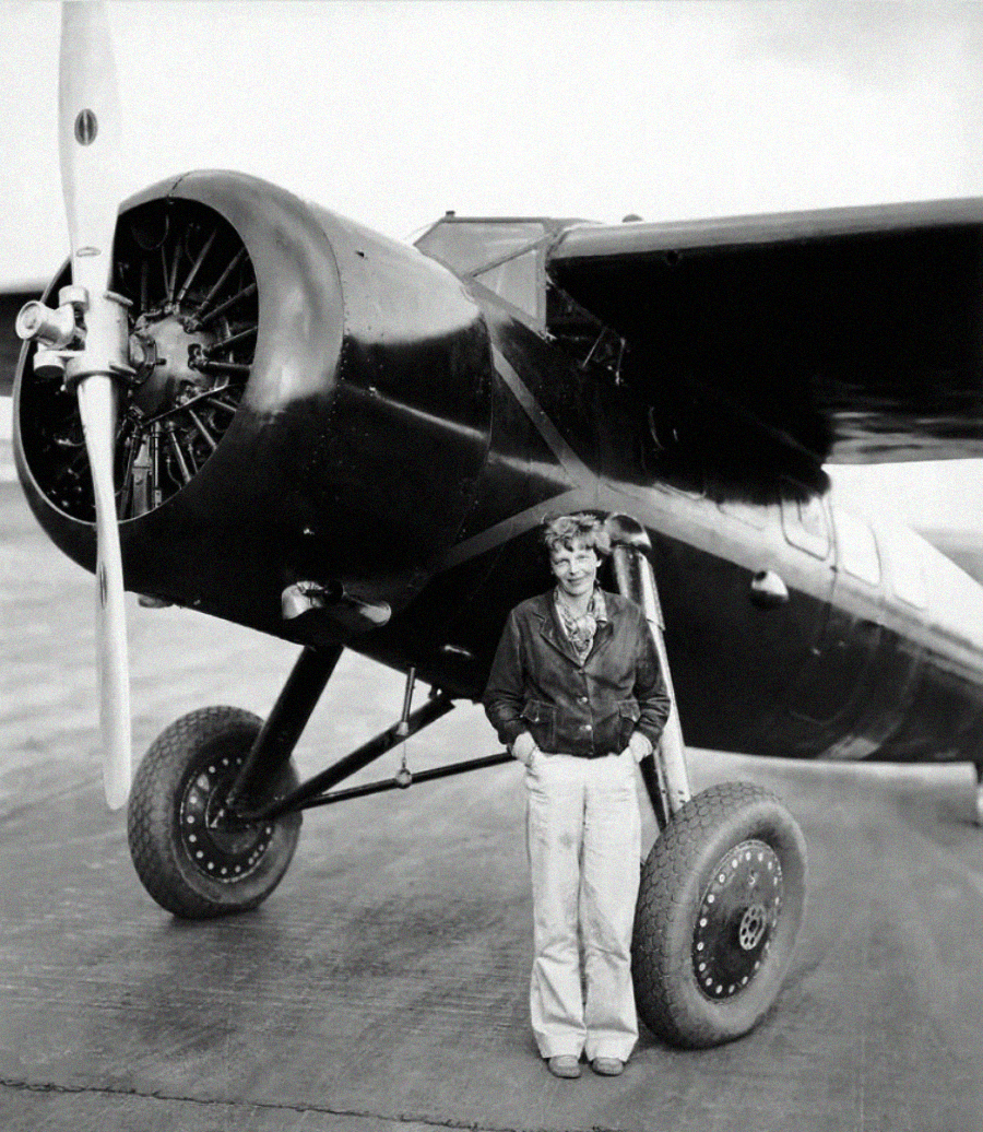 A woman in a flight jacket and trousers stands in front of a vintage single-engine propeller airplane on a runway. The plane has large wheels and a radial engine. The image is in black and white.