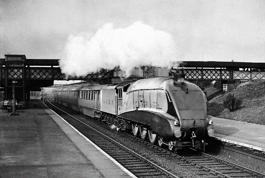 A vintage black-and-white photo of a streamlined steam locomotive pulling passenger cars, emitting smoke as it passes through a nearly empty railway station with a footbridge overhead.