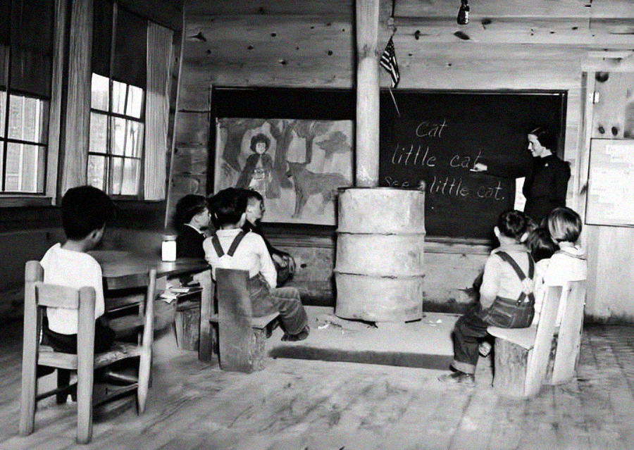 Black-and-white photo of young children sitting in a rustic classroom with wooden walls. A teacher stands near a chalkboard with the words “cat, little cat, see a little cat.” A wood stove is in the center of the room.