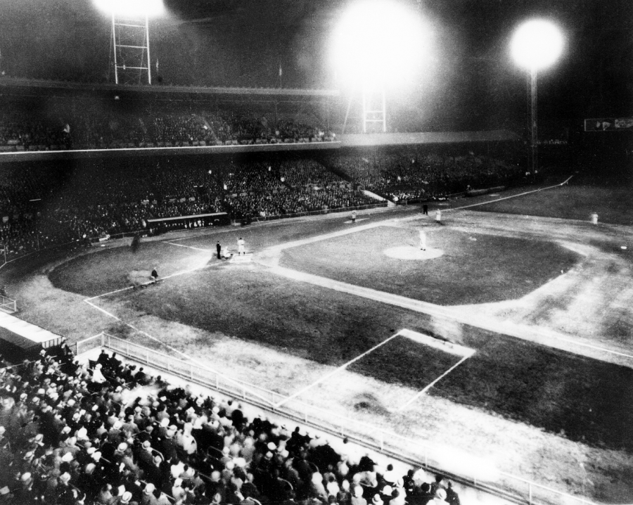 A black-and-white photo of a baseball game played at night under bright stadium lights, with a crowd of spectators filling the stands and players on the field.