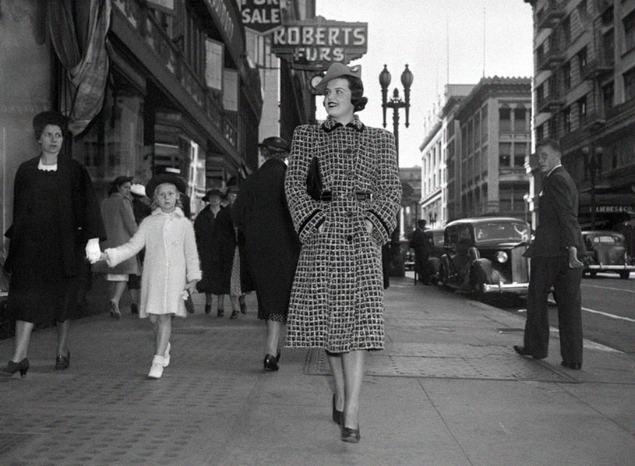 A woman in a patterned coat and hat walks confidently on a city sidewalk, surrounded by other pedestrians, vintage cars, and storefronts, including a "Roberts Furs" sign. The image appears to be from the early 20th century.