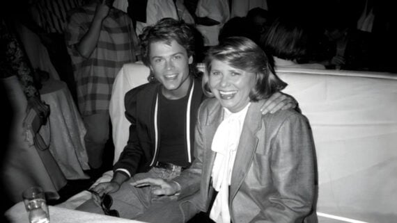 A young man and an older woman sit closely together, smiling at a table in a crowded, dimly lit room. The man has his arm around the woman, and both are dressed in formal attire.