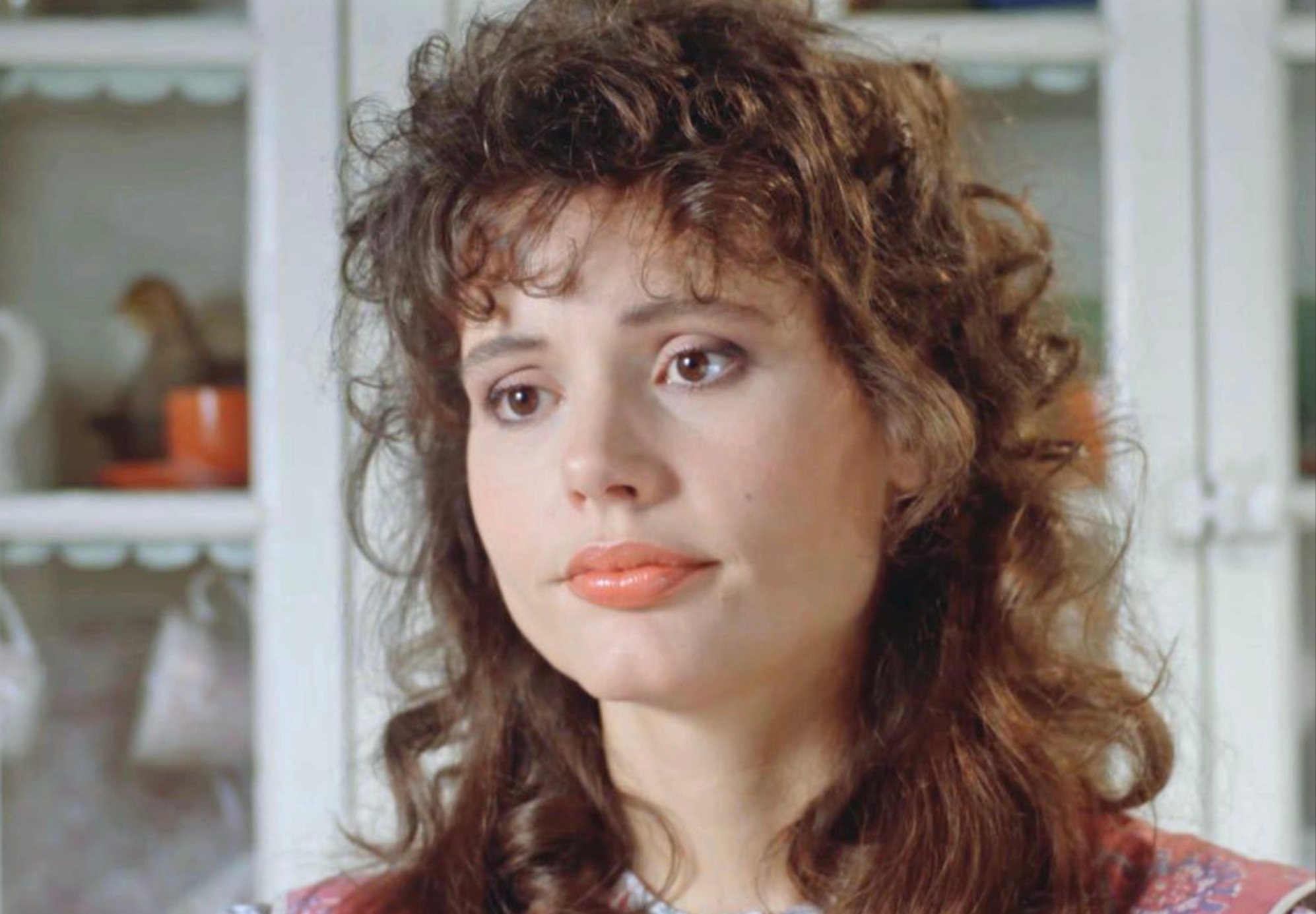 A woman with wavy brown hair and light makeup looks slightly off-camera with a neutral, thoughtful expression. She stands in front of white shelves with dishes and a cup in the background.
