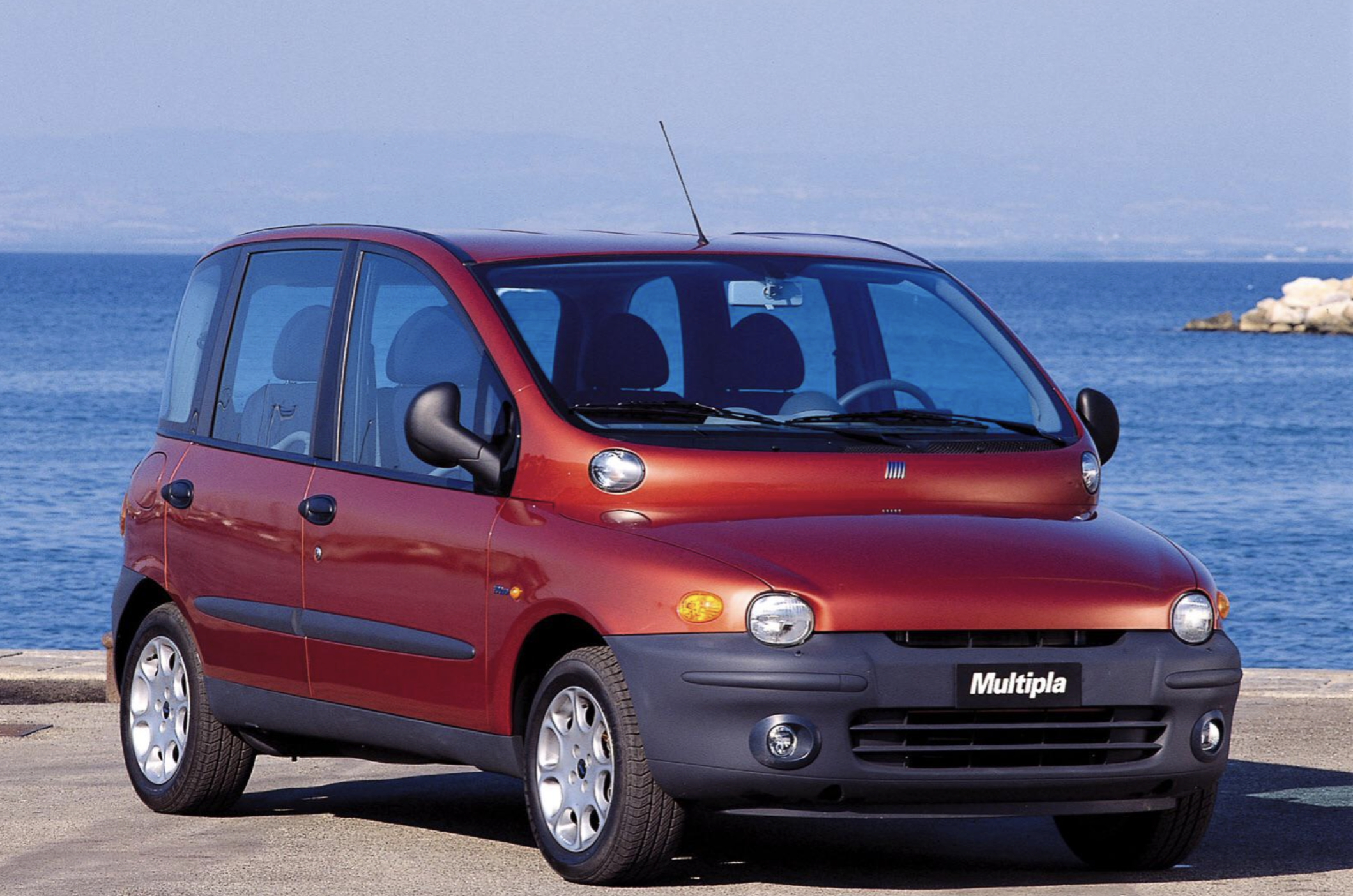 A red Fiat Multipla car is parked near a body of water with mountains visible in the background under a clear sky.