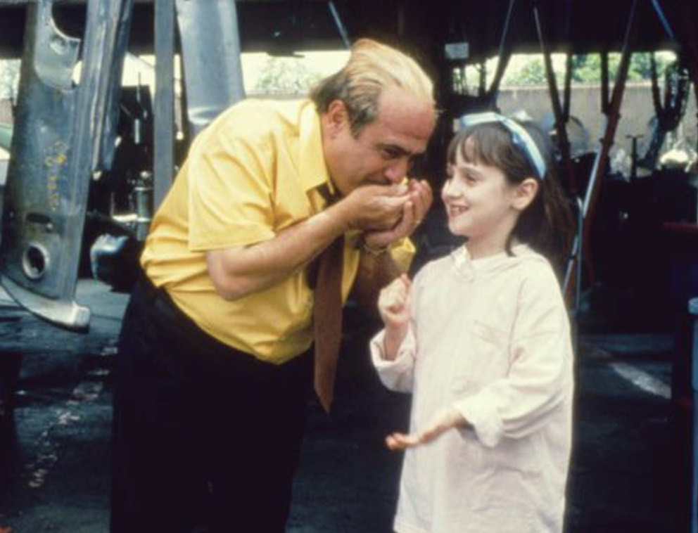 A man in a yellow shirt and tie gently holds and kisses a girl's hand. The girl, wearing a light top and blue headband, smiles. They are standing in what appears to be an industrial or workshop setting.