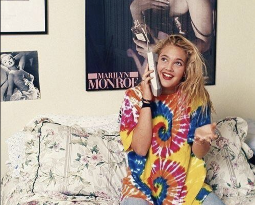 A young woman in a colorful tie-dye shirt sits on a floral bedspread, smiling while talking on a large vintage cell phone. Behind her are Marilyn Monroe posters on the wall.