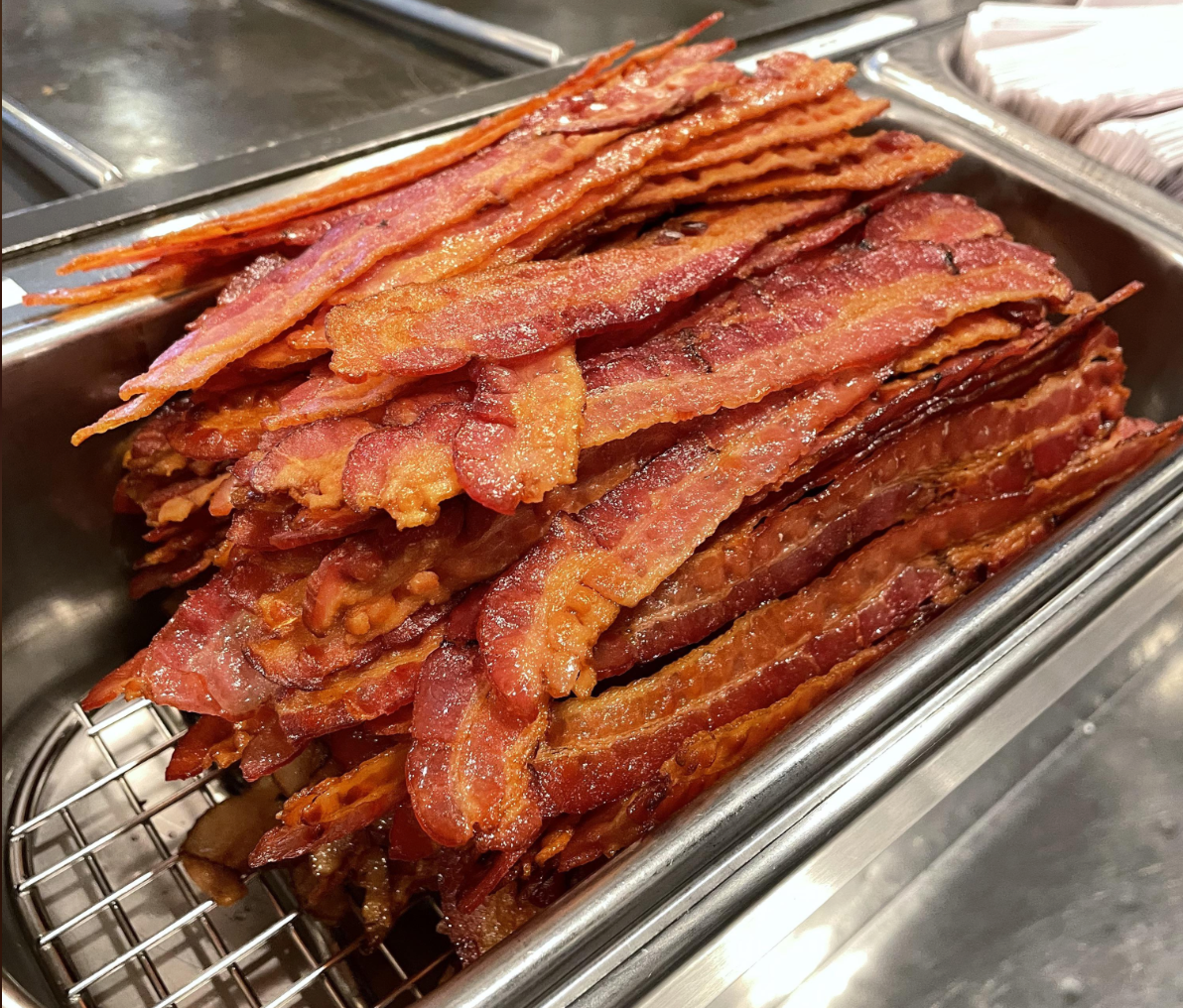 A large tray filled with crispy, cooked bacon strips stacked neatly, sitting on a metal rack in a food service setting.