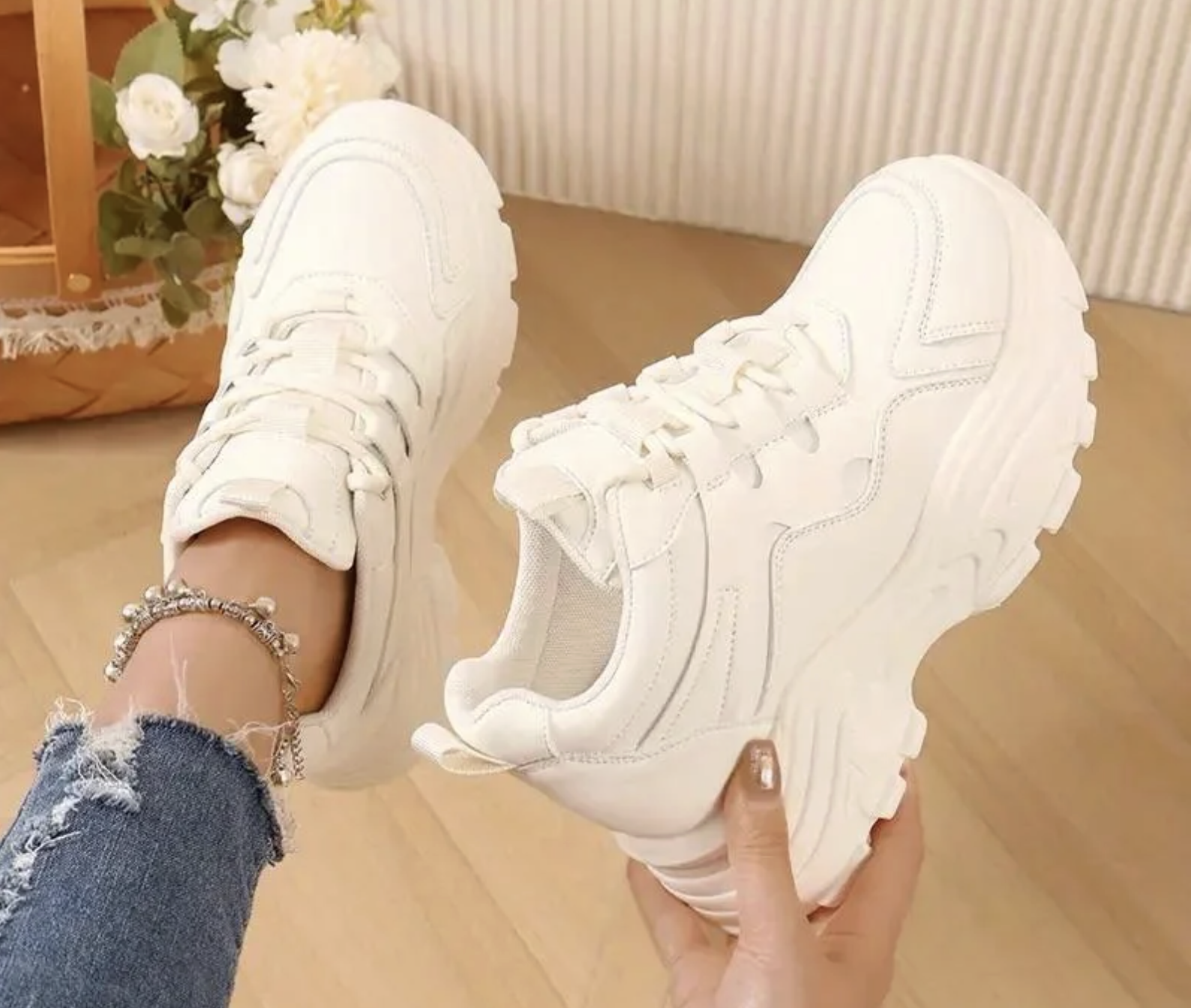 A person wearing ripped blue jeans and an anklet holds one chunky white sneaker while the other sneaker is worn on their foot. A basket and flowers are visible in the background.