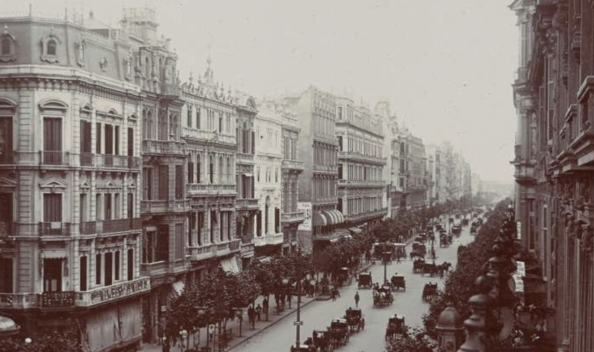 Black and white photo of a bustling city street lined with ornate multi-story buildings. Horse-drawn carriages fill the wide avenue, and trees line the sidewalks. The scene has an early 20th-century European feel.