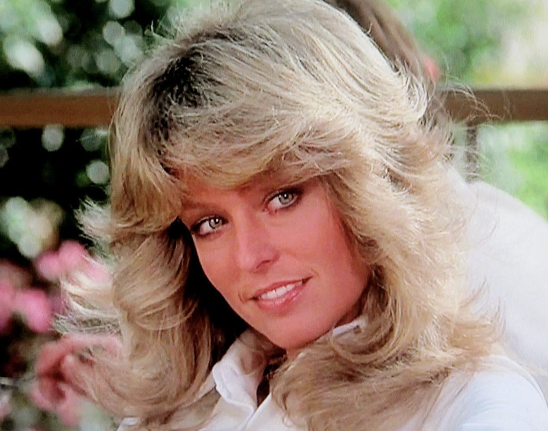 A woman with voluminous, feathered blonde hair smiles softly, wearing a white shirt. She is outdoors with a blurred background of greenery and pink flowers.