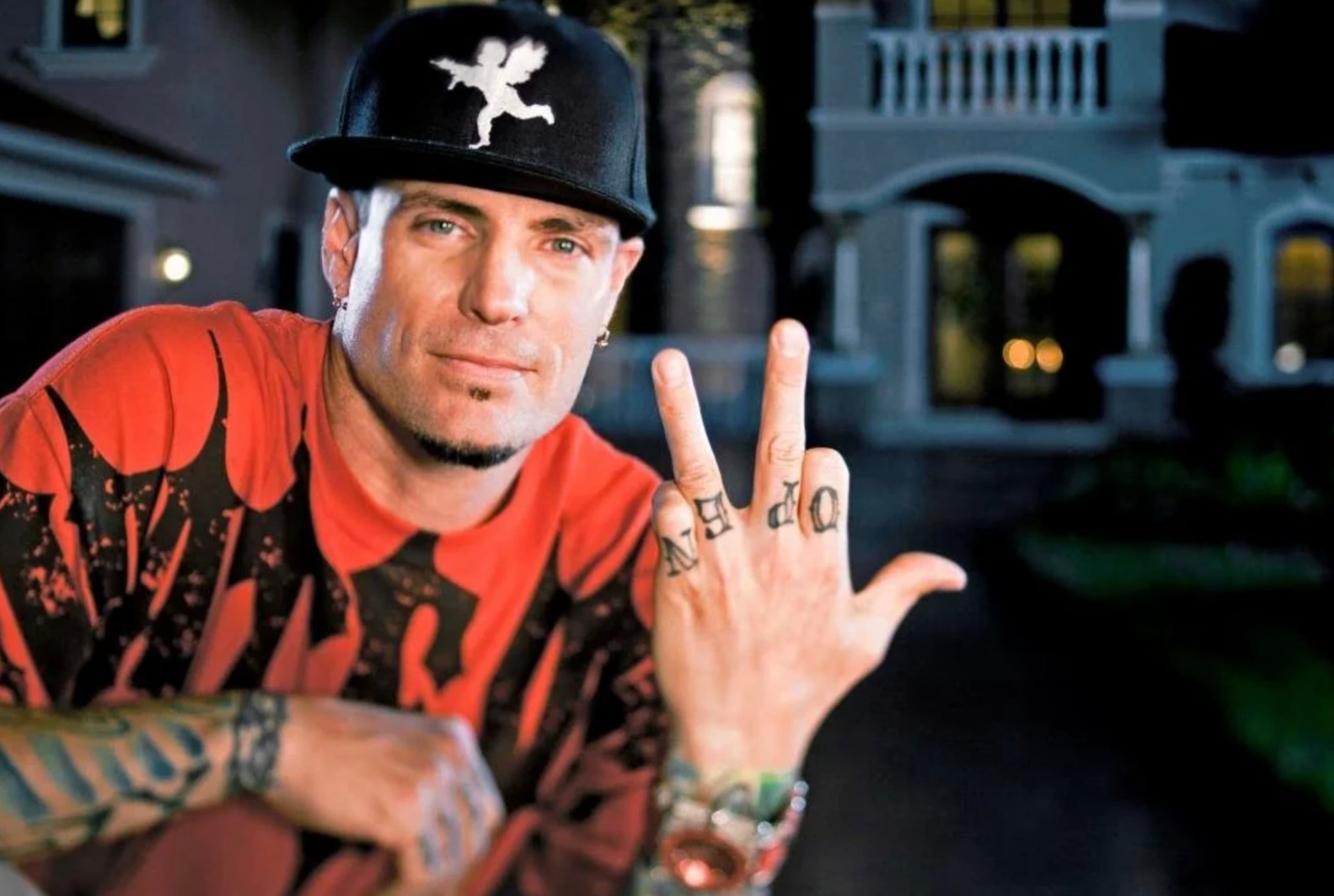 A man in a black hat, red and black shirt, and tattooed fingers poses with his hand raised, displaying the word "WILD" on his fingers, standing outside a large, well-lit house at night.