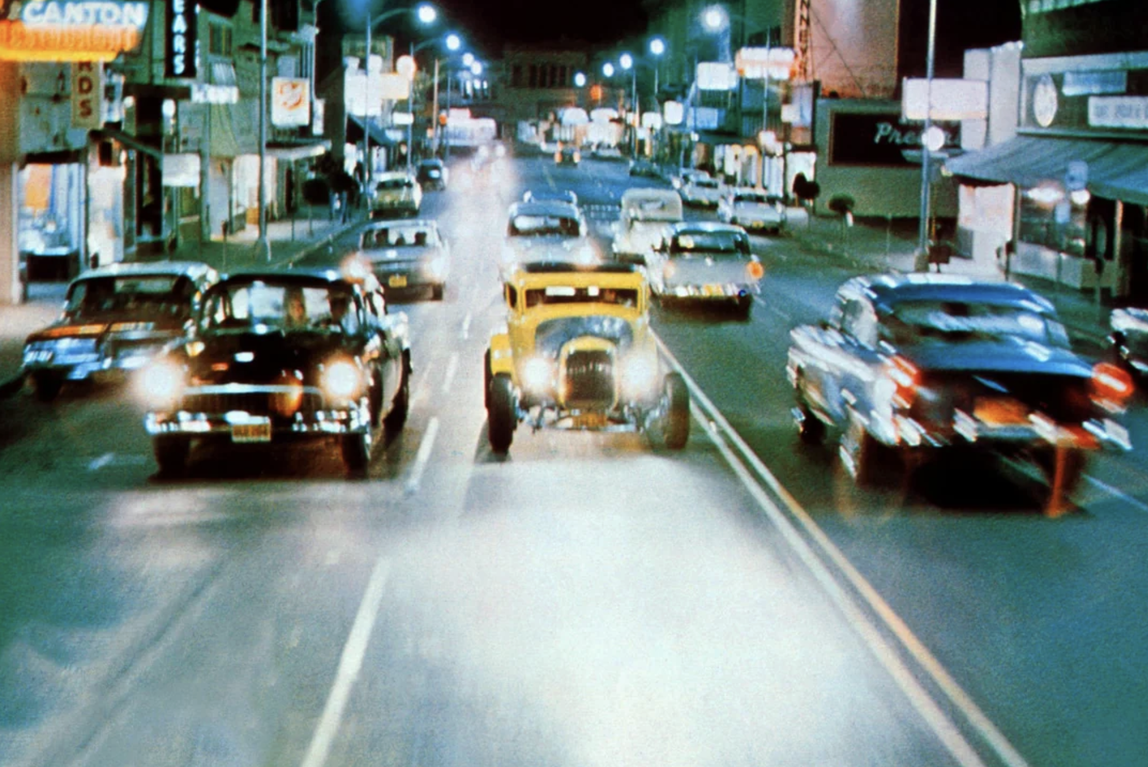 A bright yellow hot rod speeds down a lively city street at night, surrounded by classic cars under glowing streetlights and neon signs, creating a vibrant, retro atmosphere.