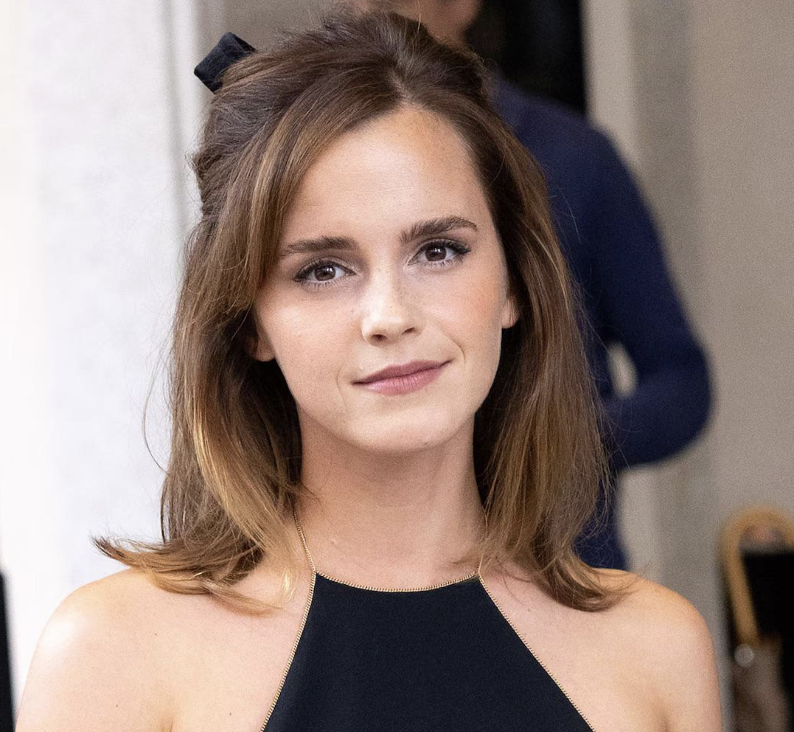 A woman with shoulder-length brown hair and a calm expression stands indoors, wearing a sleeveless black top. The background is softly blurred, suggesting an indoor setting with people and furniture.