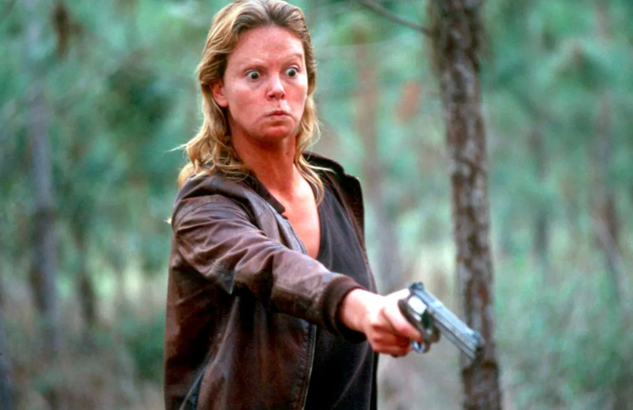 A woman with intense facial expression, wearing a brown leather jacket and dark shirt, stands outdoors in a forest, pointing a handgun forward with one arm extended. Trees and greenery are visible in the background.