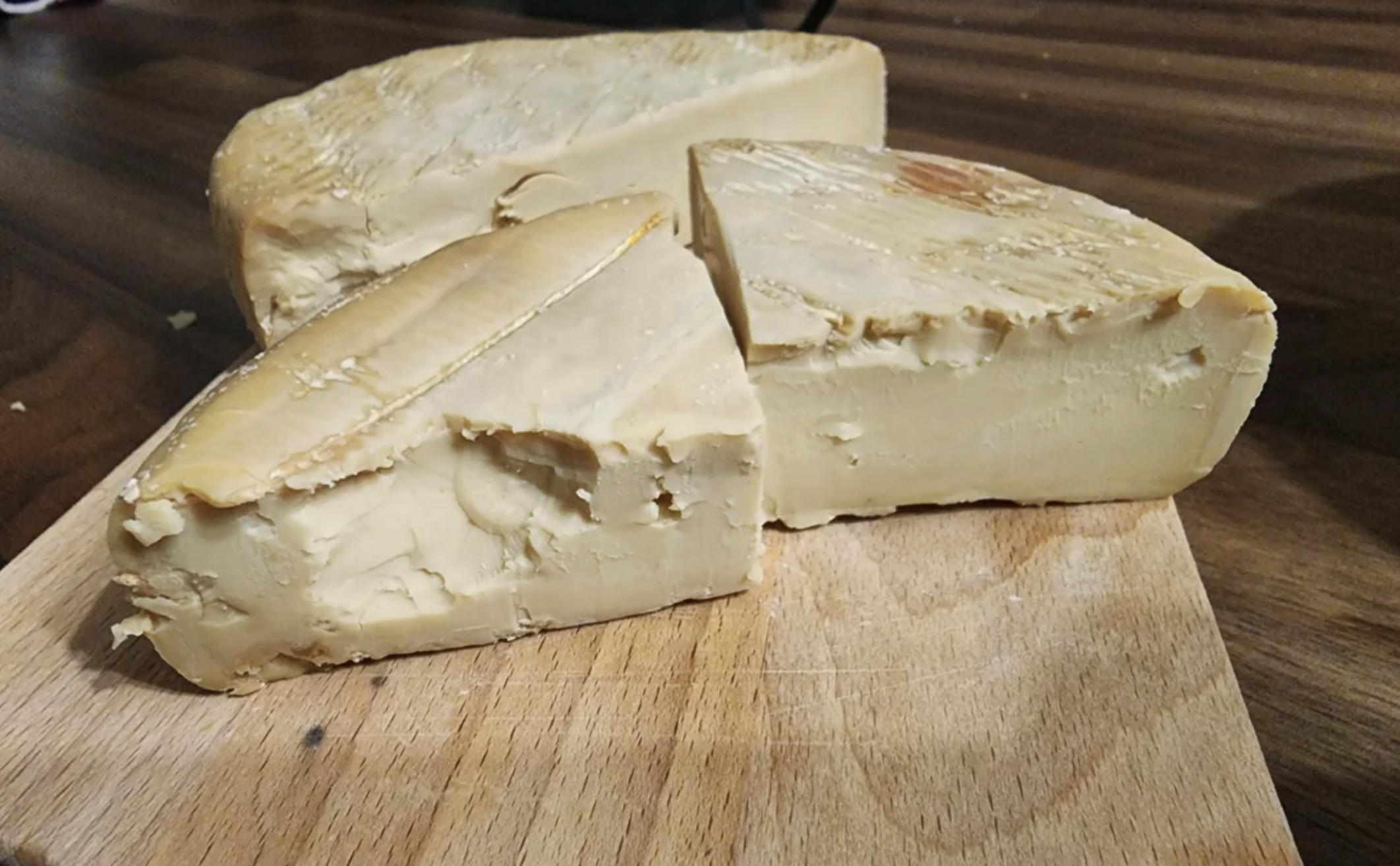 Three thick wedges of firm, beige cheese rest on a wooden cutting board. The cheese has a slightly crumbly texture and an uneven, natural rind. The surface appears creamy with some cracks and indentations.